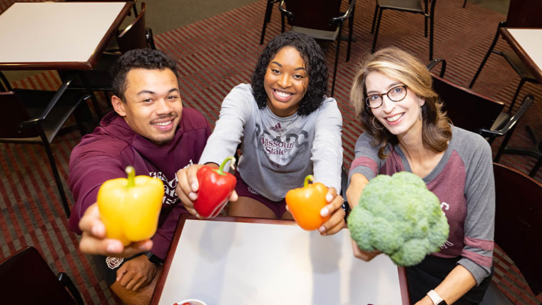 Two students and their instructor hold up bell peppers (of various color) and broccoli.