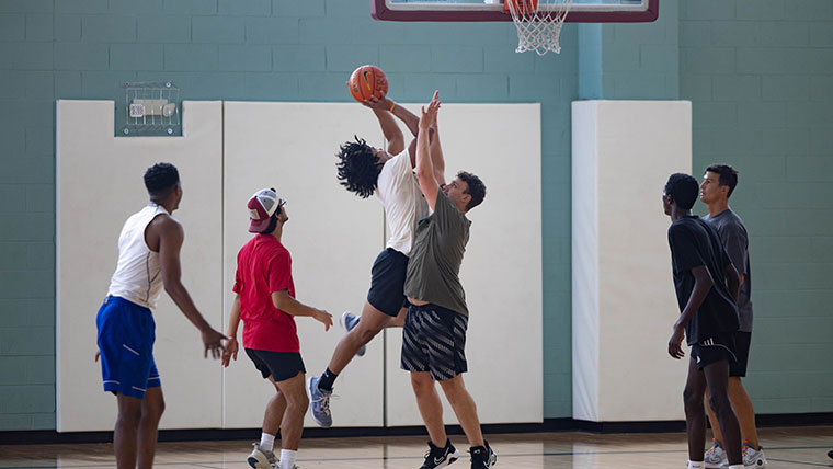 A group of six guys take their basketball skills to the court. One player jumps and prepares for a shot as another player tries to block him.